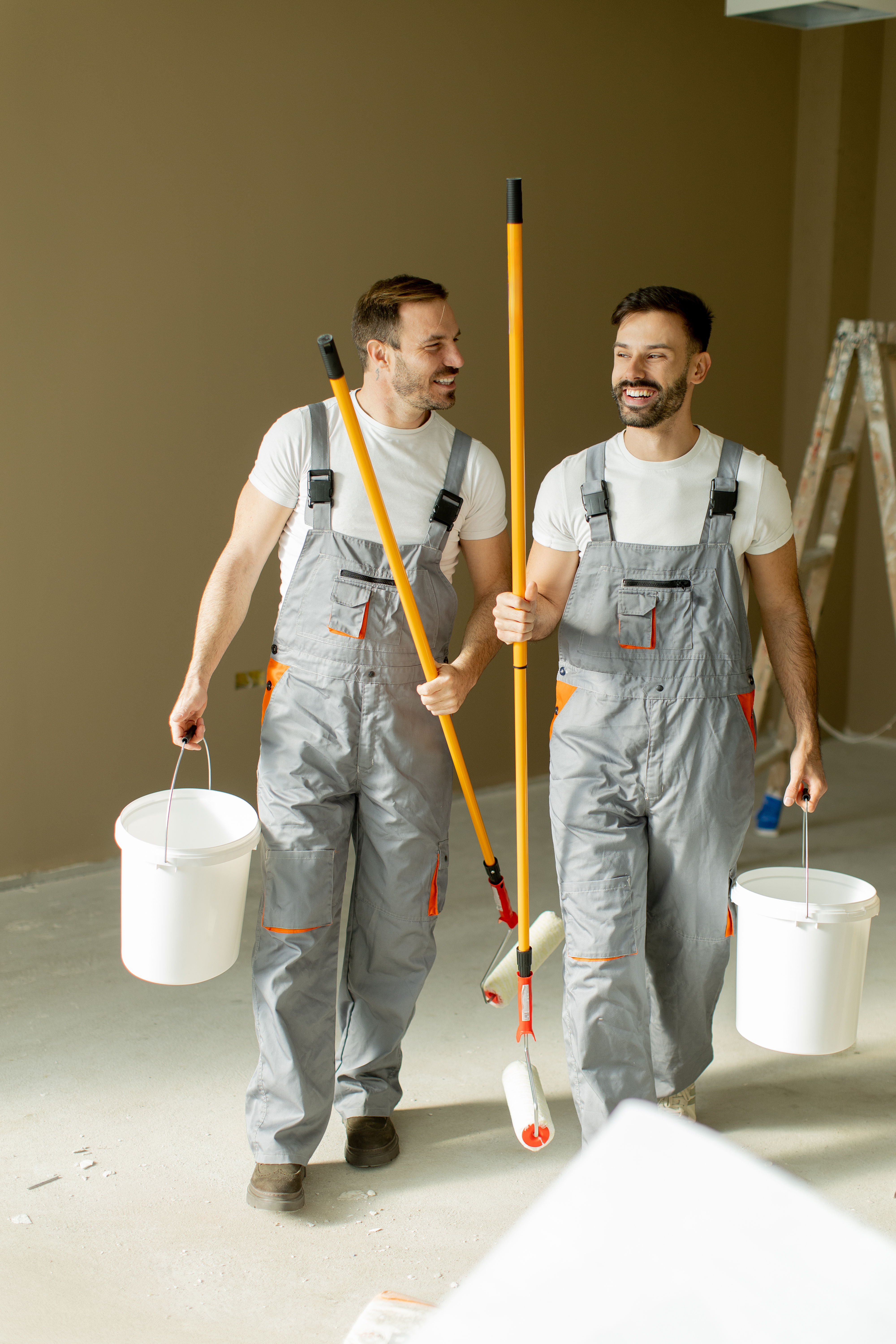 Two smiling men in gray overalls walk through a room that is being painted. They carry paint buckets and long paint rollers, ready to start their work in the freshly renovated space.